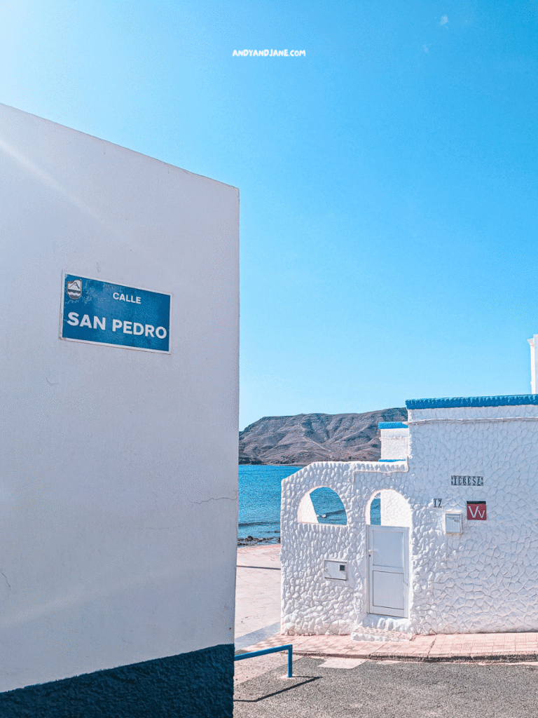A white building on Calle San Pedro in Las Playitas, Fuerteventura faces the sea, with a mountainous coastline visible in the background under a clear blue sky.
