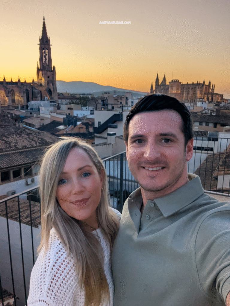 A couple poses for a selfie on a rooftop at sunset, with a backdrop of historic buildings and a serene sky in warm hues.