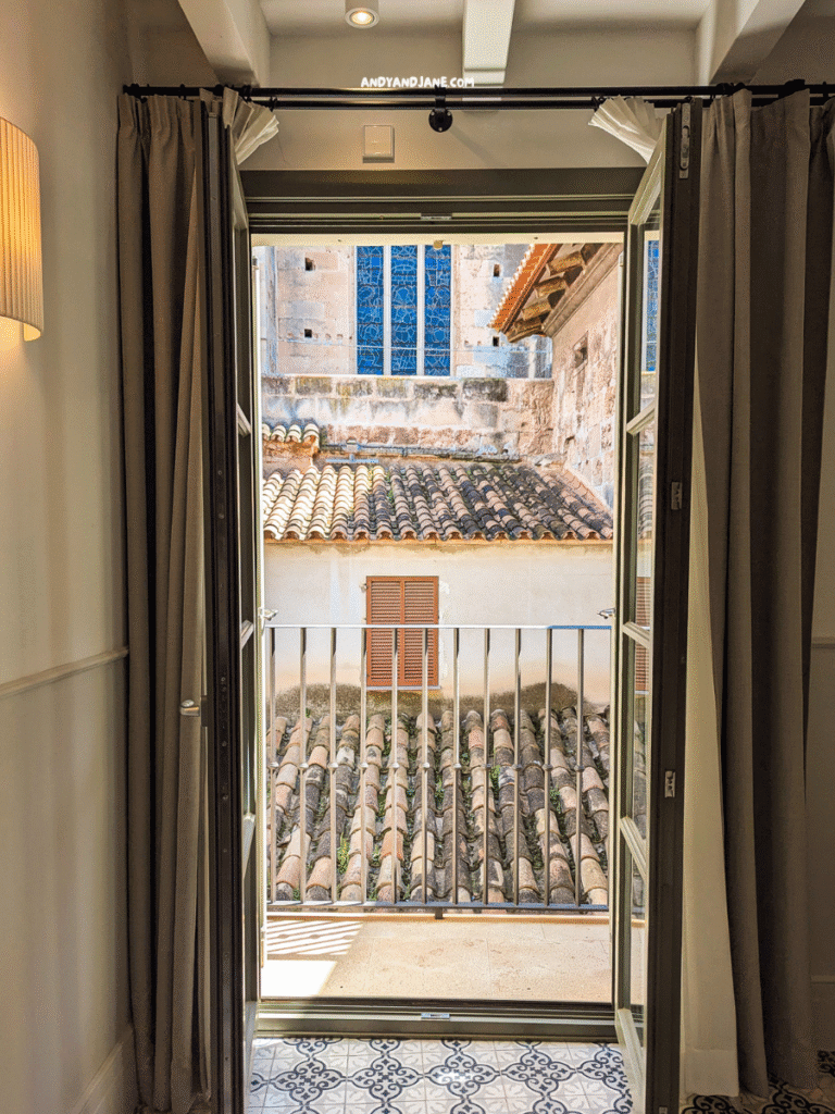 A view from an open balcony overlooking tiled rooftops and a building with blue stained glass windows. Soft curtains frame the scene.
