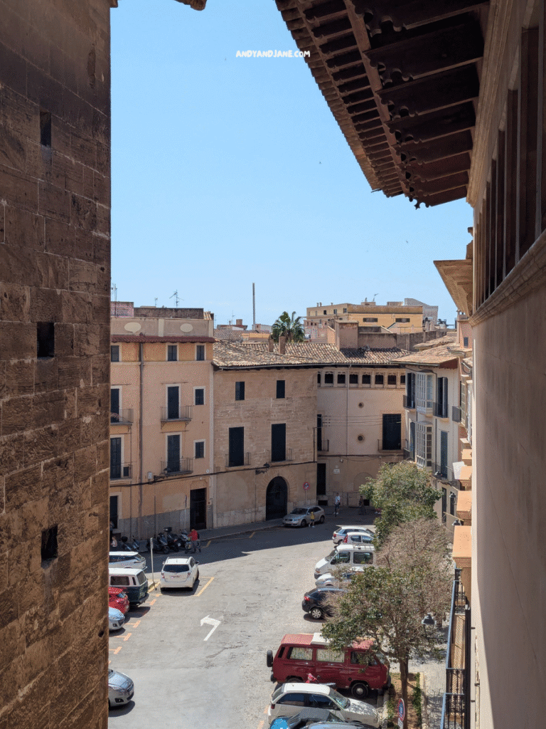 View of a sunny square surrounded by historic buildings, with parked cars and a hint of palm trees against a clear blue sky.