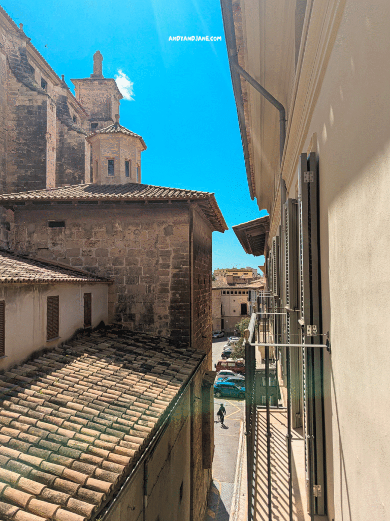 View from a balcony showing narrow cobblestone street, adjacent buildings with tiled roofs, and a clear blue sky.