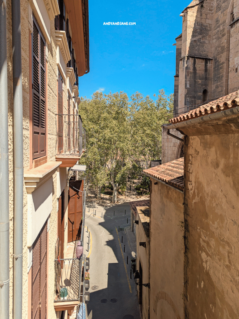 View from a balcony overlooking a narrow street, lined with historic buildings and trees under a clear blue sky.