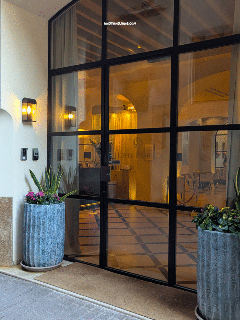 The entrance of Hotel Basilica, Palma featuring large glass doors, warm lighting, and potted plants flanking the doorway.