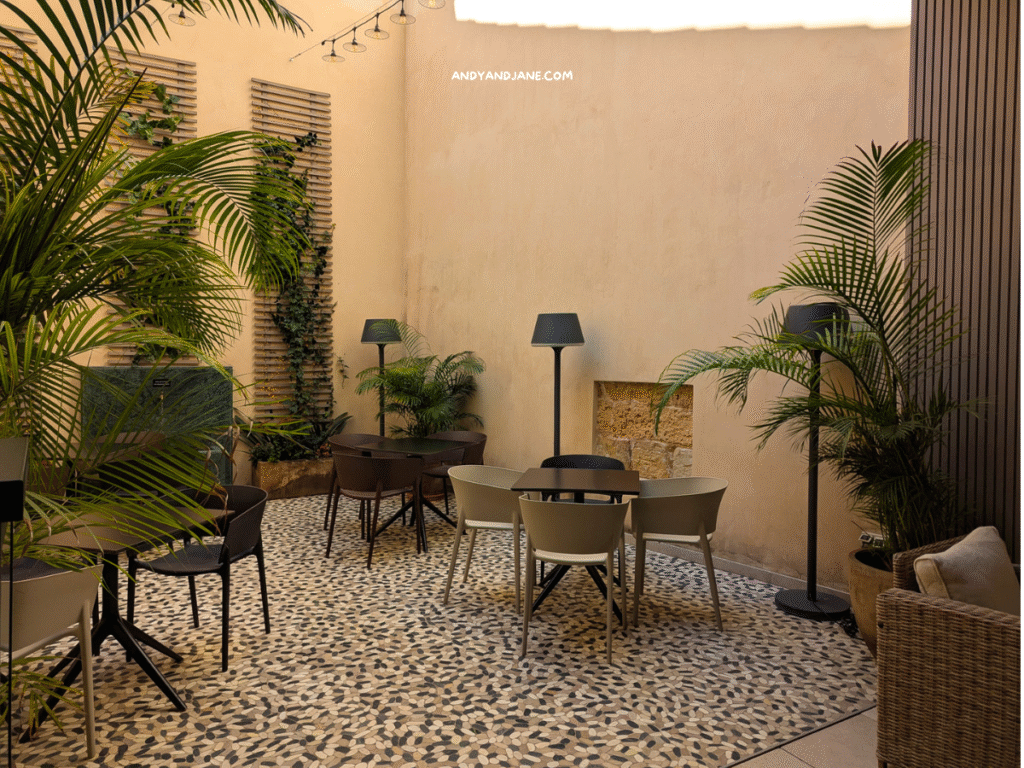 A serene outdoor seating area featuring stylish chairs, lamps, lush greenery, and a patterned stone floor, surrounded by warm, neutral walls.