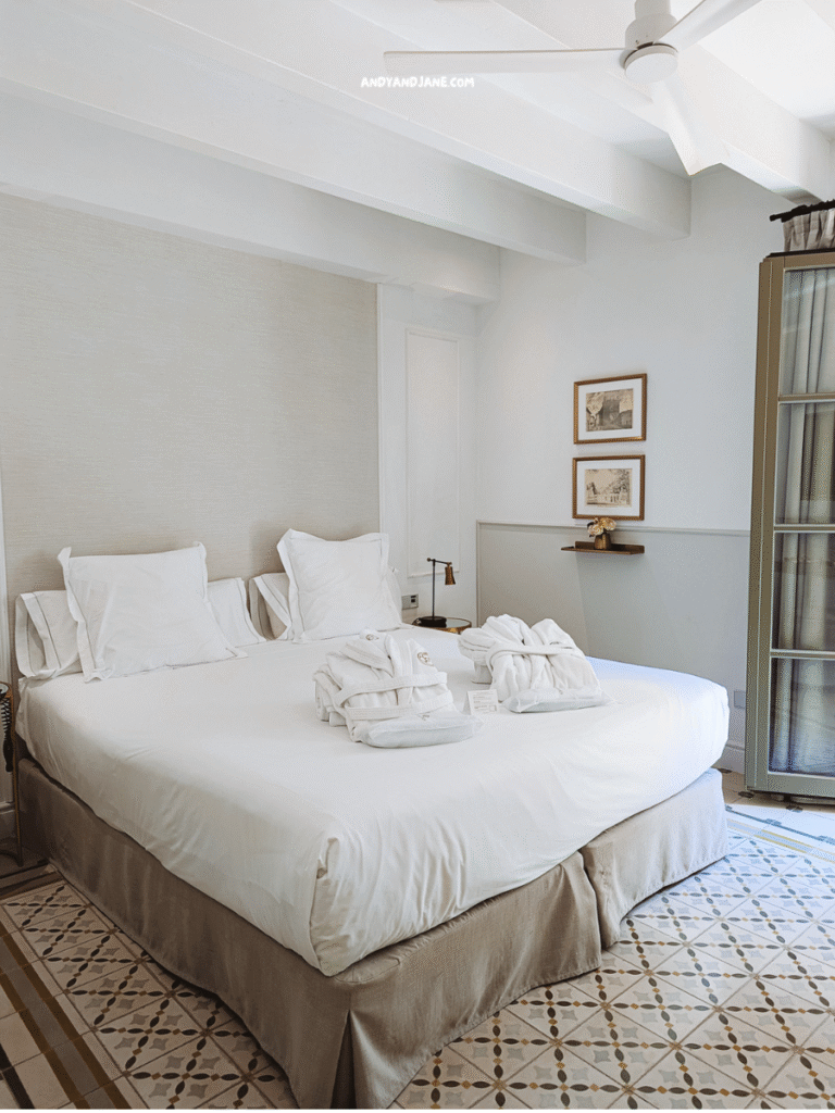 A cozy bedroom at Hotel Basilica featuring a neatly made double bed, plush white towels, and artistic decor, illuminated by natural light.