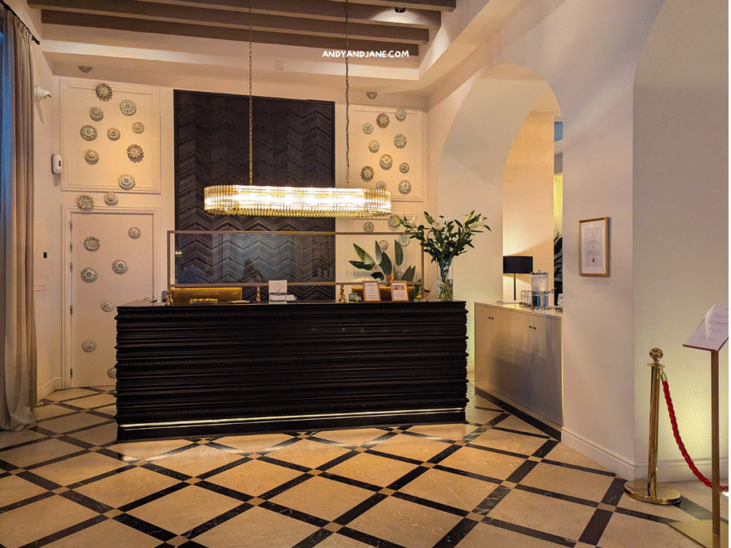 Elegant hotel reception area at Hotel Basilica with a dark wooden desk, chandelier, decorative wall plates, and a welcoming floral arrangement.
