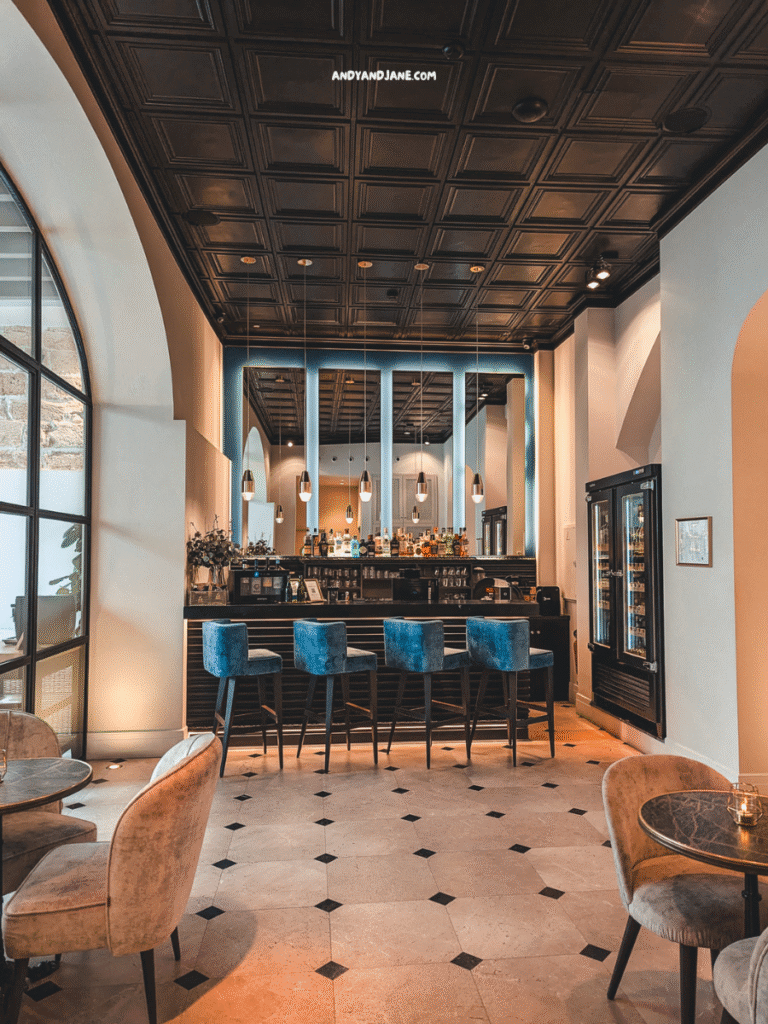 Modern bar in Hotel Basilica with blue velvet stools, elegantly designed ceiling, and a chic ambiance, featuring marble tables in the foreground.