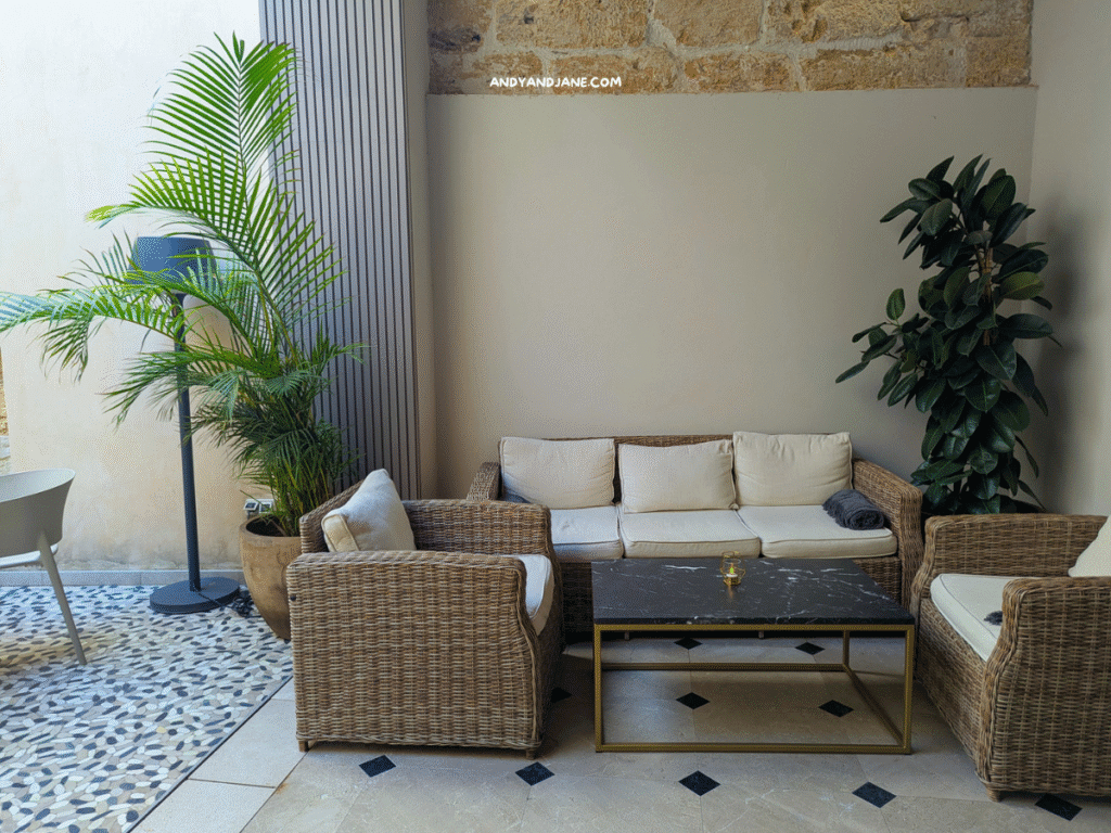 A cozy patio setting with rattan furniture, a marble coffee table, and lush greenery against a stone wall backdrop.