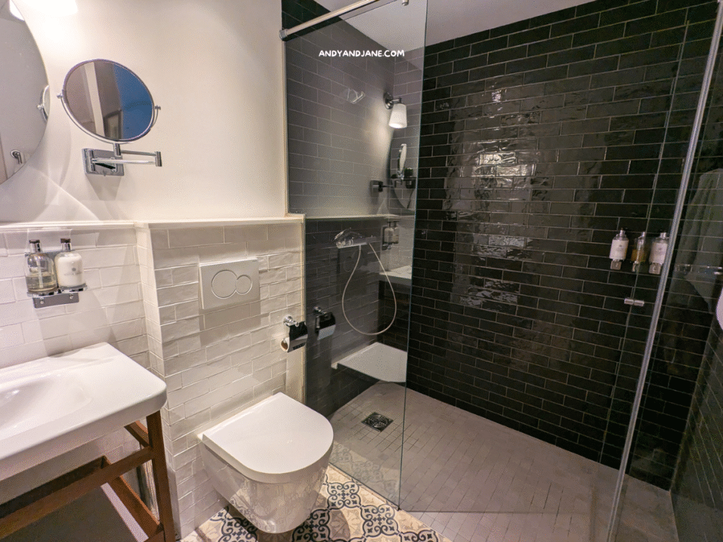 Modern bathroom featuring a white sink, wall-mounted mirror, shower area with dark tiles, and patterned floor tiles.