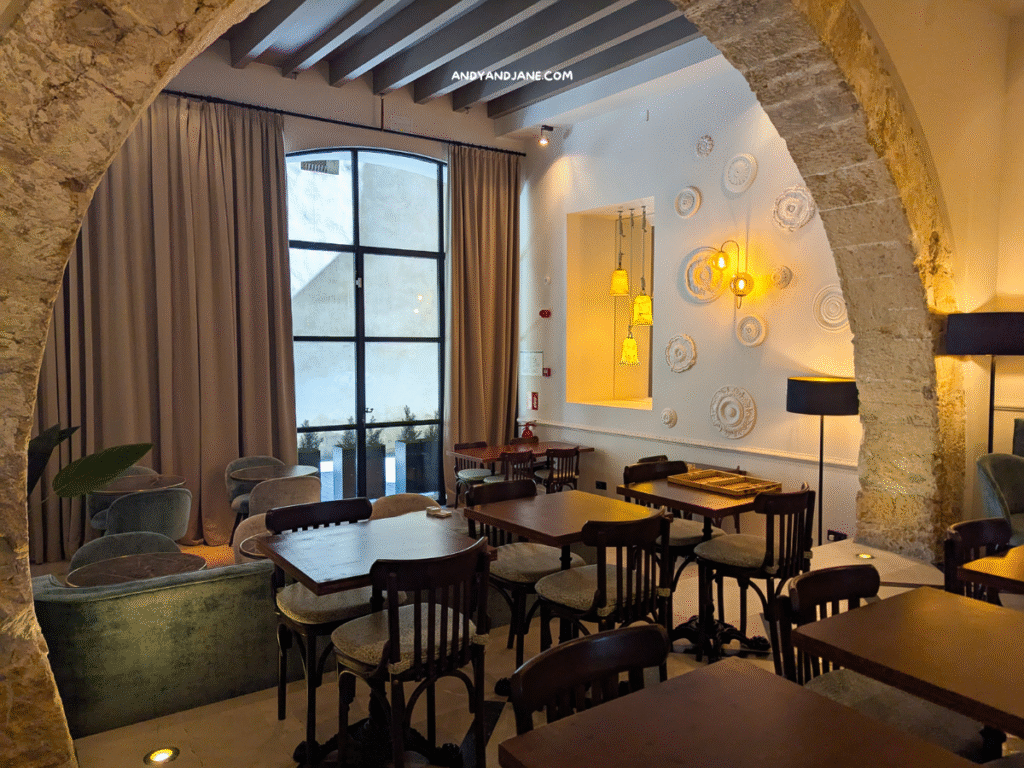 Restaurant area in hotel basilica featuring wooden tables, plush seating, decorative wall art, and warm lighting through large windows and curtains.