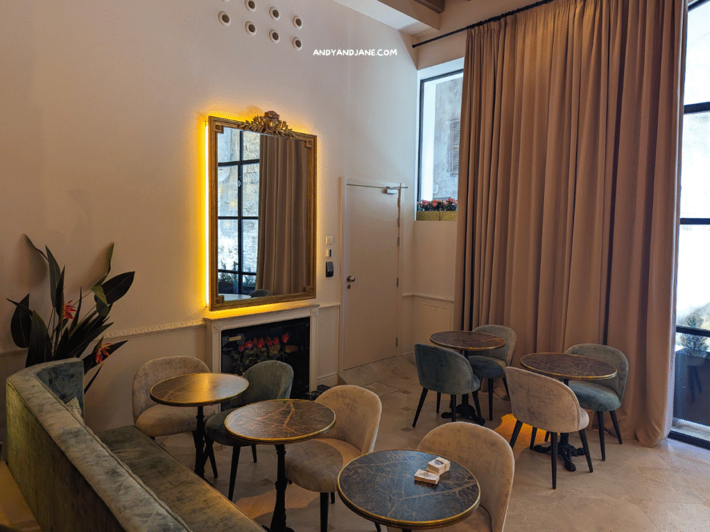 Cozy restaurant at Hotel Basilica featuring plush seating, marble-topped tables, a large illuminated mirror, and soft curtains by a window.