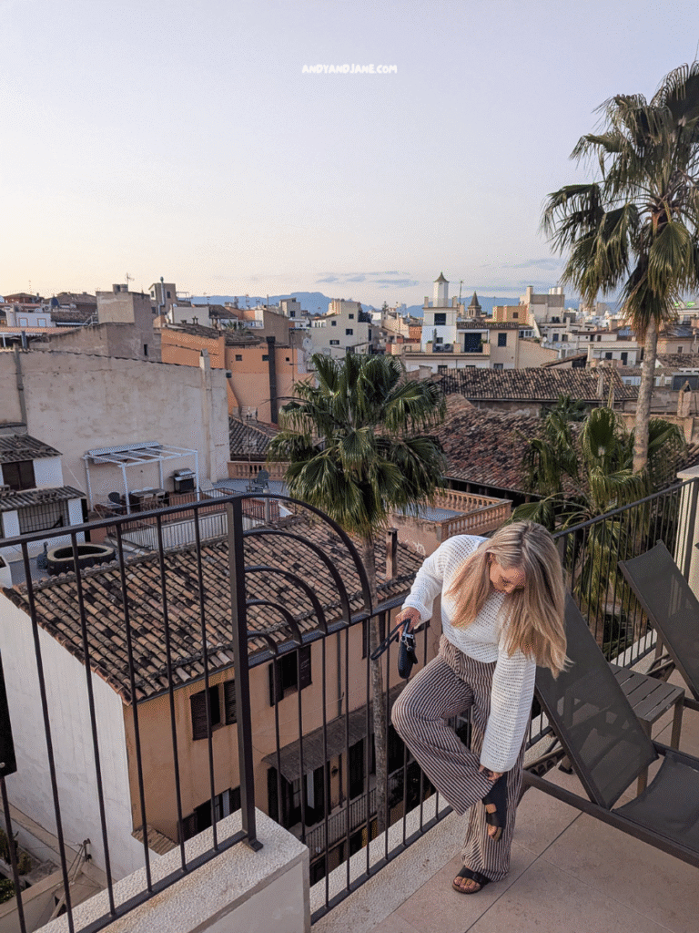 A woman adjusts her outfit while standing on a balcony overlooking a scenic cityscape with palm trees and rooftops at sunset.