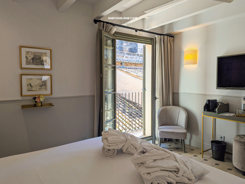 A cozy hotel room featuring a bed with rolled towels, an open balcony, framed artwork, a chair, and a flat-screen TV.