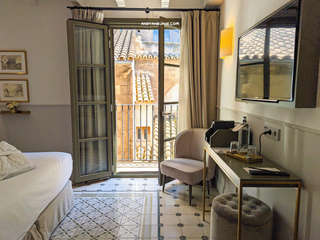 Hotel Basilica room with a bed, balcony doors, TV, elegant chair, and patterned tile floor, offering a view of terracotta rooftops.