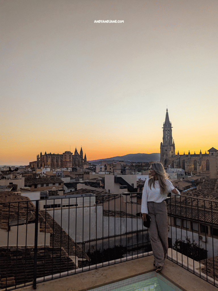 A person stands on a balcony overlooking a city at sunset, with historic buildings and mountains in the background.