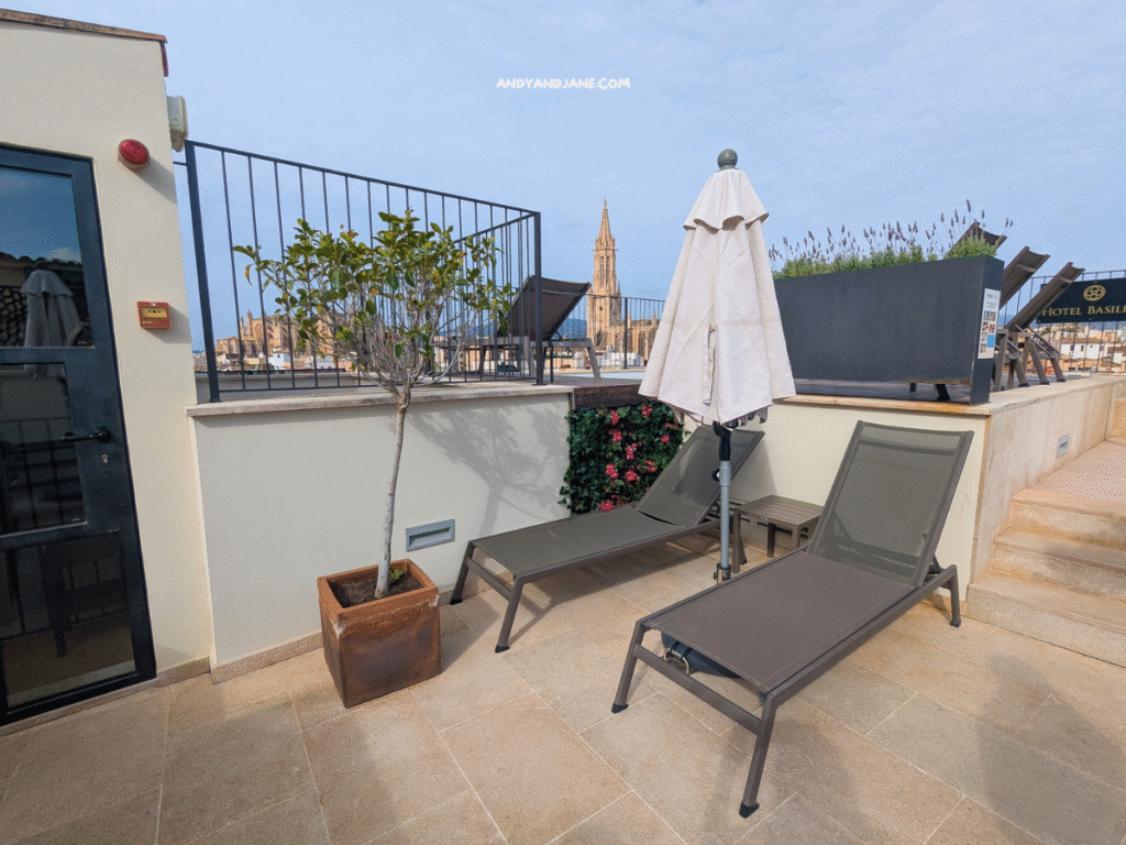 Rooftop terrace with two loungers, an umbrella, potted plants, and a view of a cathedral tower against a cloudy sky.
