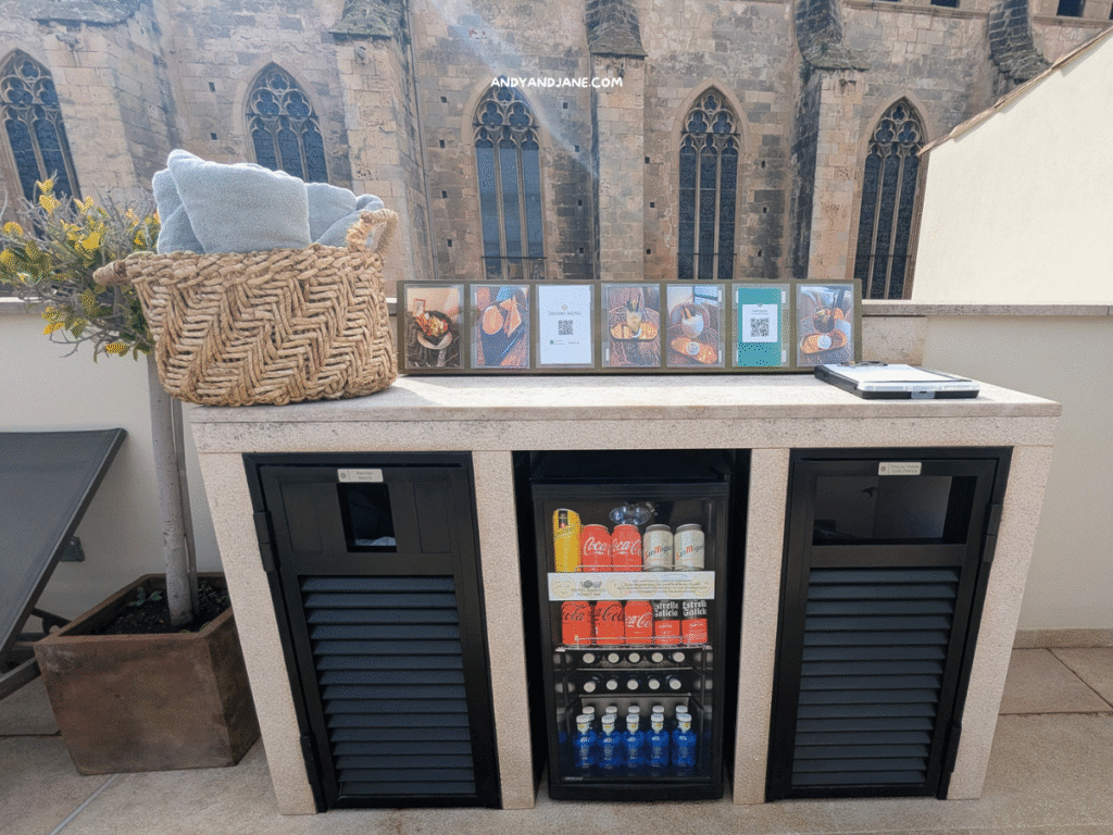 A stylish outdoor bar area with a wicker basket of towels, beverage coolers, and framed pictures against a historic stone building backdrop.