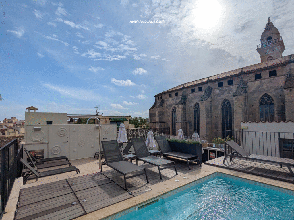 Rooftop pool area at Hotel Basilica features lounge chairs and umbrellas with a historic church in the background under a partly cloudy sky.