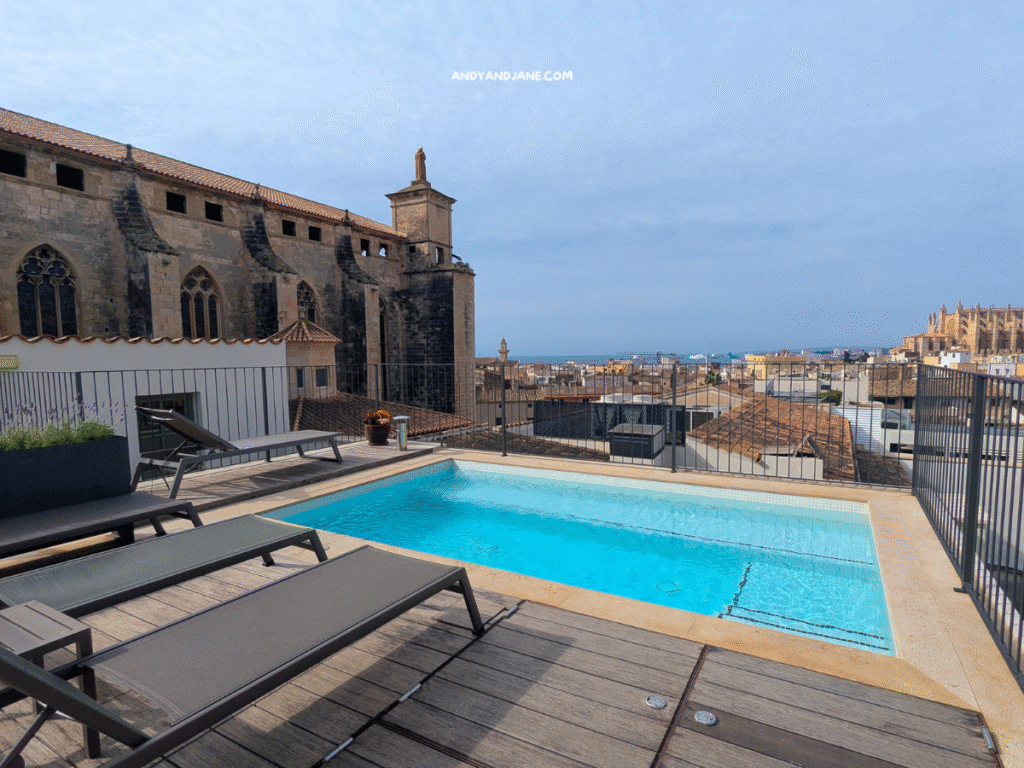 Rooftop pool at Hotel Basilica with lounge chairs, overlooking historic architecture and the sea, under a cloudy sky.