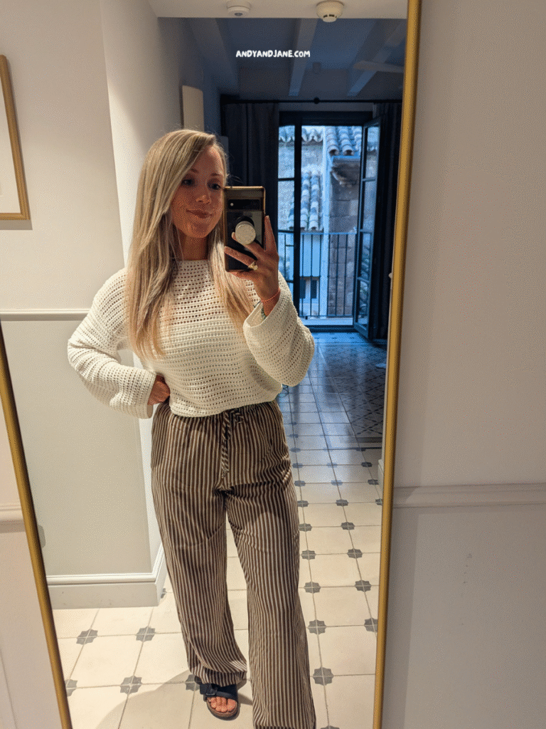 A person stands in front of a mirror, wearing a cozy white sweater and striped wide-leg pants, with a balcony visible in the background.