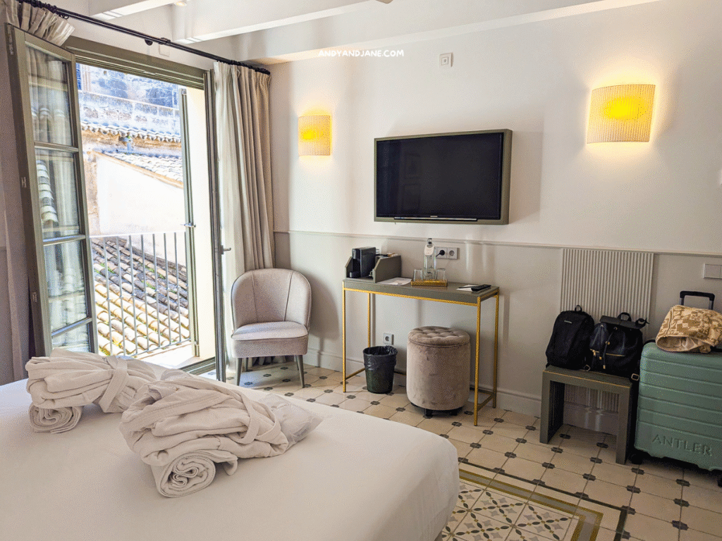 A cozy colonial room at Hotel Basilica featuring a bed with rolled towels, a wall-mounted TV, a chair, luggage, and a window with views of rooftops.