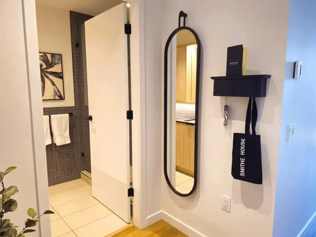 Contemporary hallway with a sleek oval mirror, small black shelf holding books, and a hanging tote. Open door shows bathroom with towels. Modern decor.