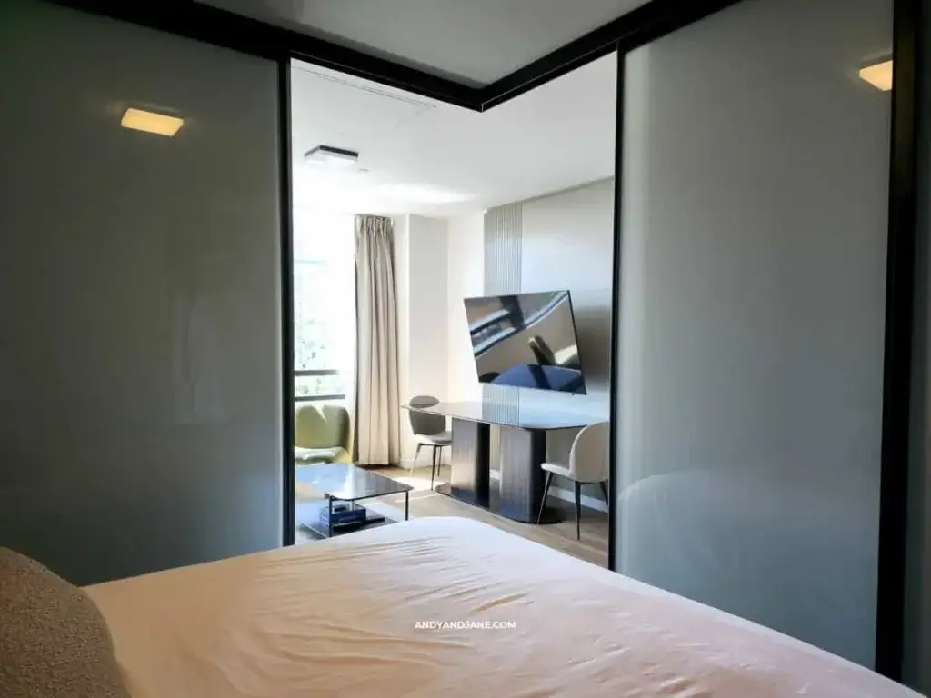 Modern hotel room with a bed in the foreground, sliding frosted glass doors, a flat-screen TV, and a seating area with two chairs and a table. Neutral tones.