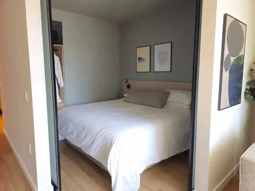 A neatly made bed with white bedding sits against a light blue accent wall in this Smithe House bedroom, flanked by two framed artworks; closet visible to the left, light wood floor throughout.