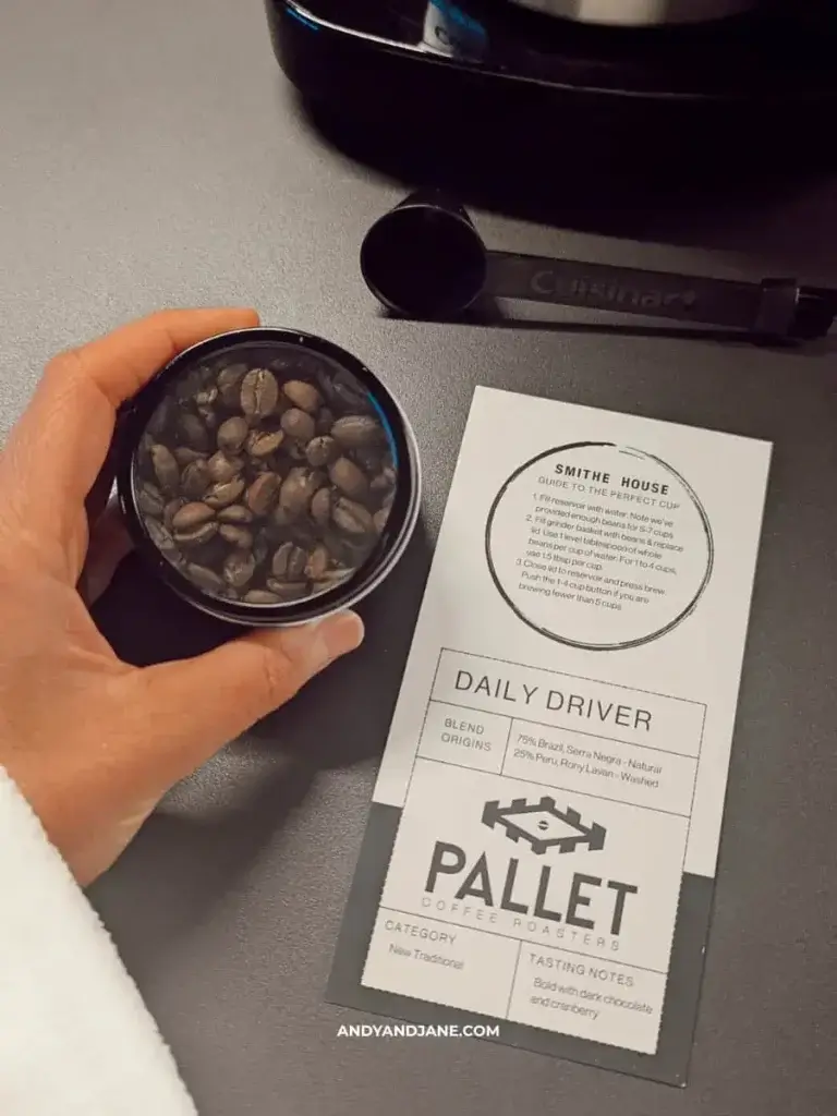 A hand holds a clear container of coffee beans beside a tasting note card from Pallet Coffee Roasters, detailing blend and brewing tips.