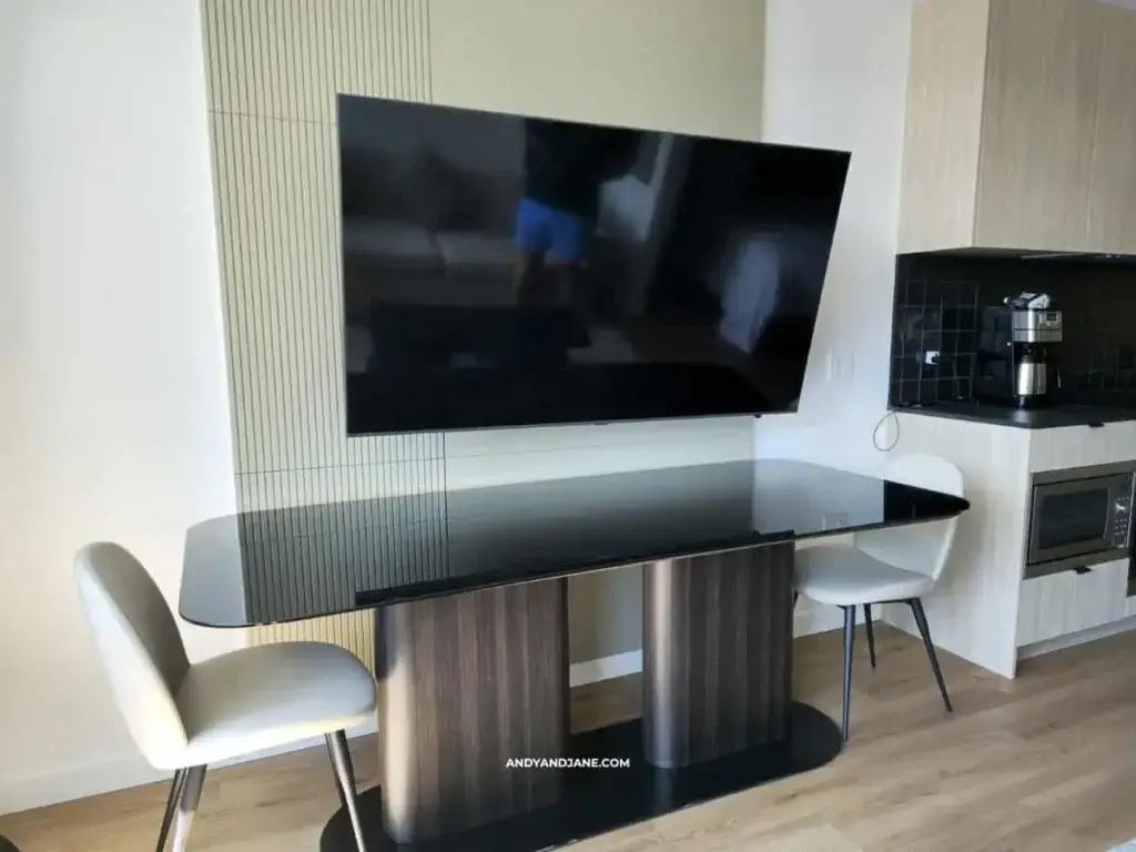 A modern dining setup featuring a sleek black table, two white chairs, and a large wall-mounted TV above a minimalist kitchen area.