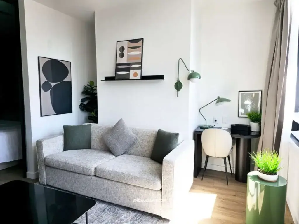 Modern living room with a grey sofa, abstract art, and green accents. A small desk with a lamp and chair sits by a bright window, creating a calm workspace.