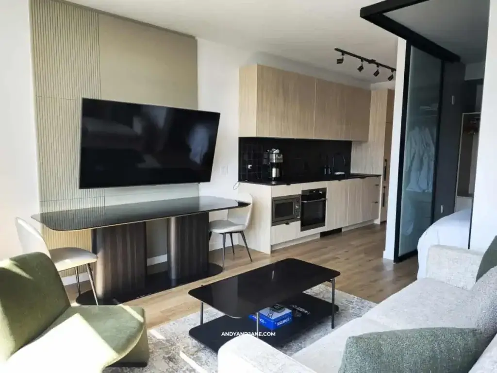 Modern apartment interior at Smithe House with a sleek design. Features a wall-mounted TV, compact dining area with two chairs, minimalist kitchen, and cozy sitting area.