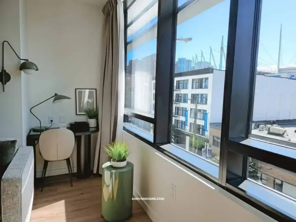 Bright corner of a modern room with a small desk, chair, and lamp by tall windows overlooking city buildings. A plant adds a touch of nature.