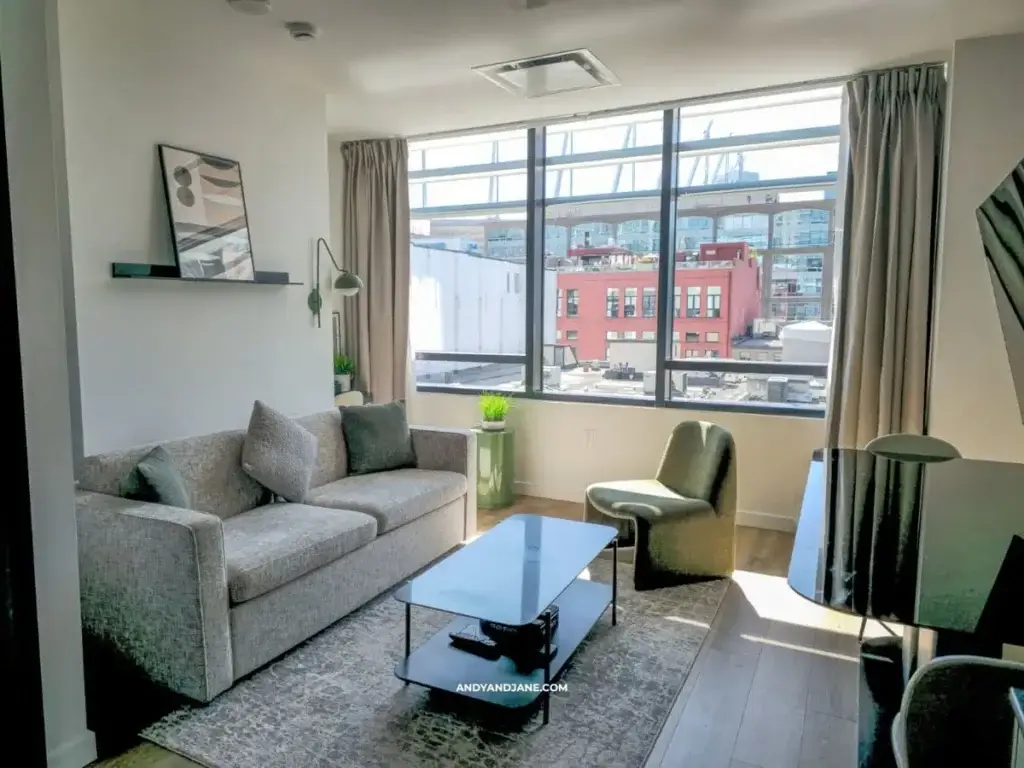 Modern, sunlit living room with gray sofa, green chair, and glass coffee table on a rug. Large window reveals cityscape with colorful buildings.