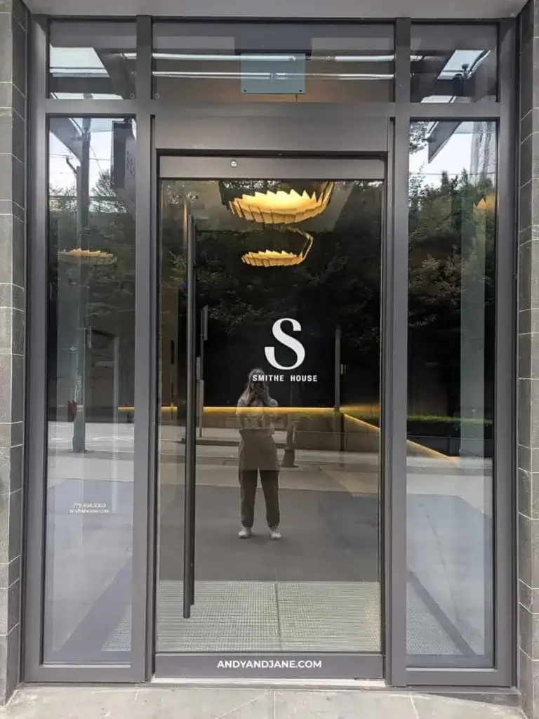 Reflection of a person taking a photo is visible in the glass door of Smithe House. The sleek, modern doorway suggests a refined setting.