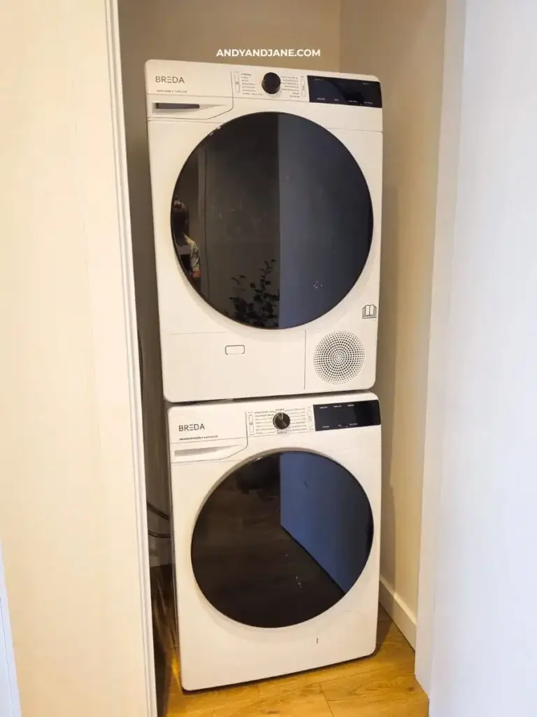 Stacked white washing machine and dryer in a narrow laundry nook, with sleek black control panels. Warm wooden floor complements the clean setup.
