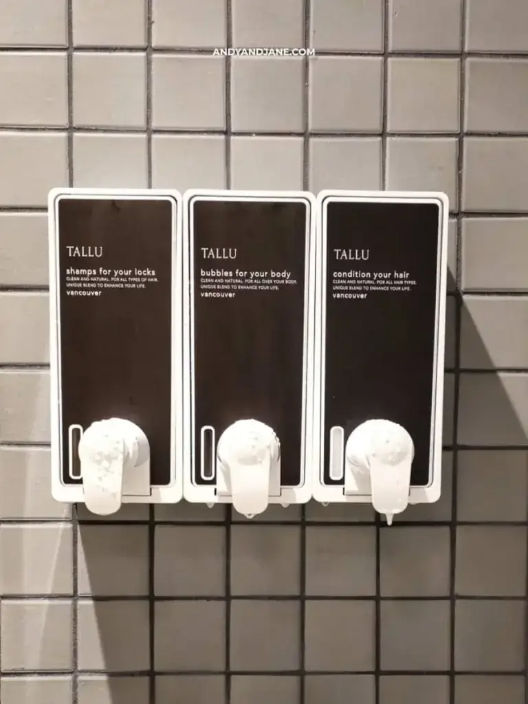 Three wall-mounted dispensers with white pumps on gray tiles. Labels read "shampoo," "body wash," and "conditioner." Minimalist and tidy setup.