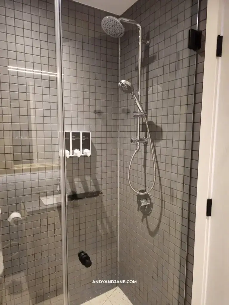 A modern shower with gray square tiles, featuring a rain showerhead, handheld nozzle, and three wall-mounted soap dispensers. Glass door adds elegance.