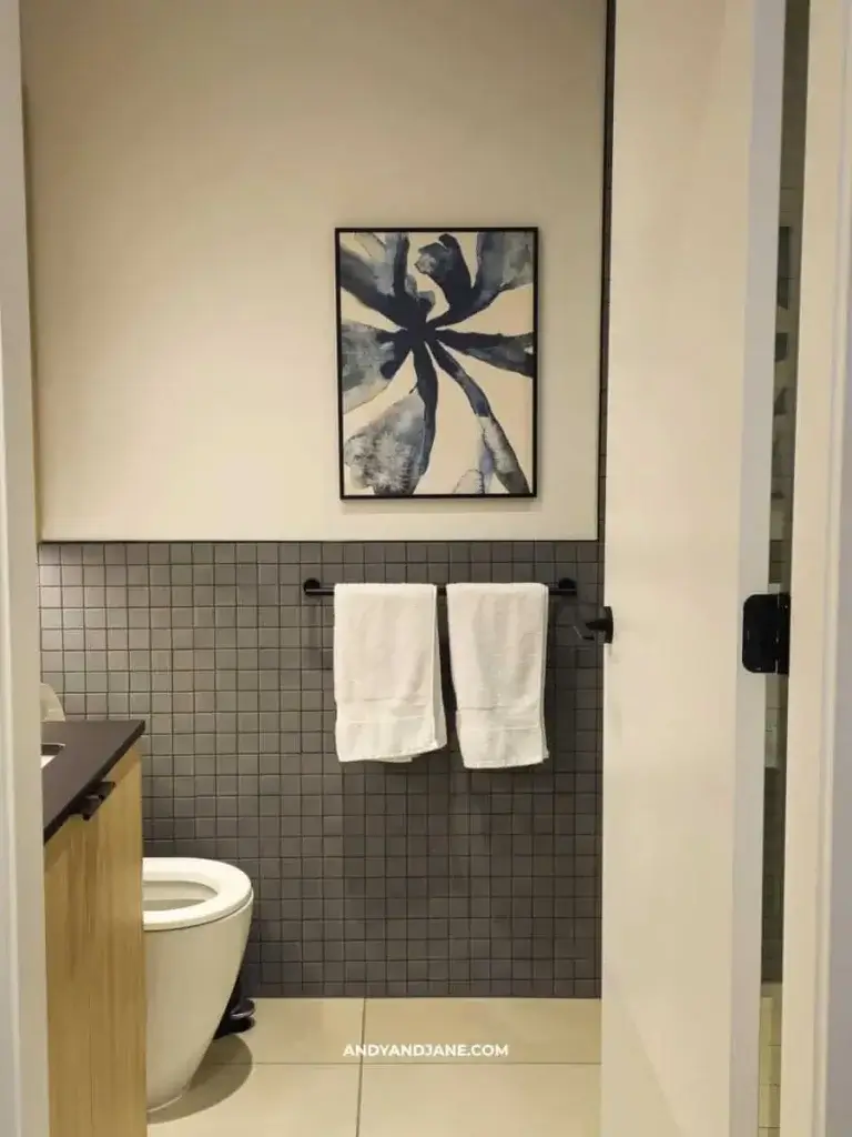 Modern bathroom with a minimalist design. Features a gray tiled wall, abstract blue and white artwork, white towels, and a wooden vanity next to a toilet.