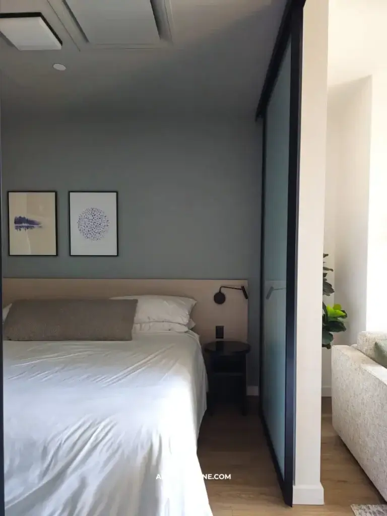 Cozy bedroom featuring a bed with white linens, a dark nightstand, framed art on the wall, and a sliding glass partition.