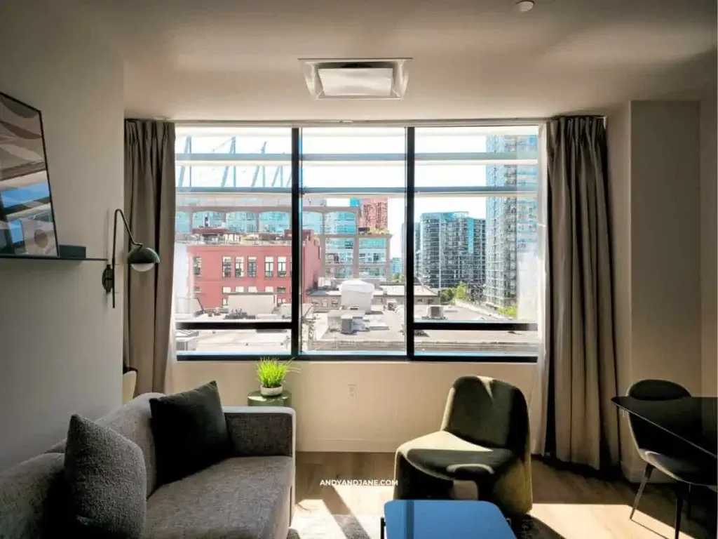 Bright, modern living room with a gray sofa, green plant, and large windows showcasing a vibrant cityscape and buildings outside.