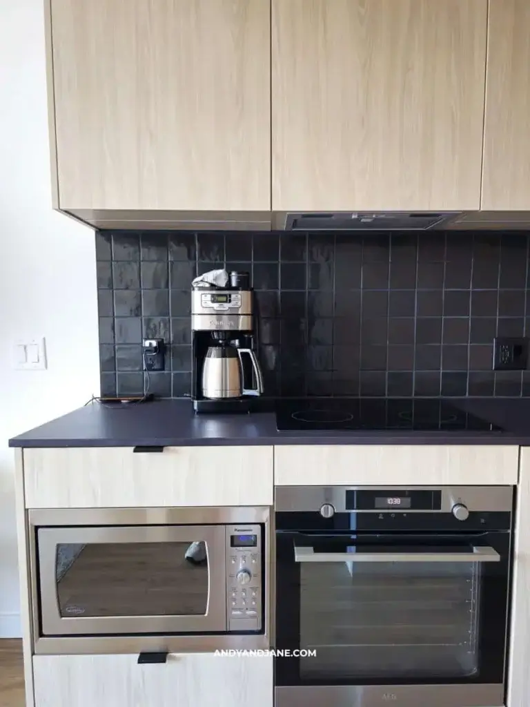 Modern kitchen with a wood cabinet, black tiled backsplash, coffee maker, microwave, and an oven. Clean, sleek, and minimalist design.
