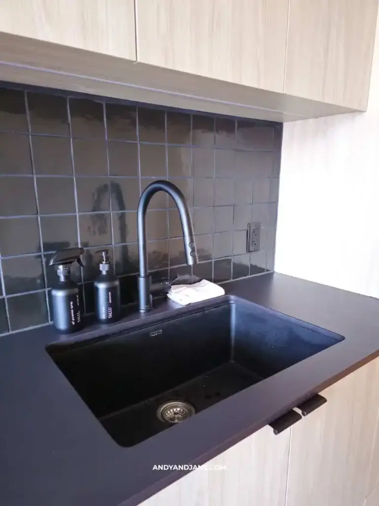 Modern kitchen sink with matte black fixtures, a dark countertop, and glossy black tiled backsplash. Soap dispensers and sponge nearby. Minimalist design.