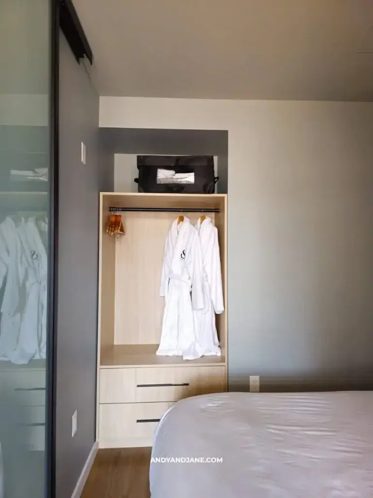 Open closet at Smithe House with two white bathrobes on hangers, a black bag on the top shelf, and two drawers below, situated beside a bed in a modern room.