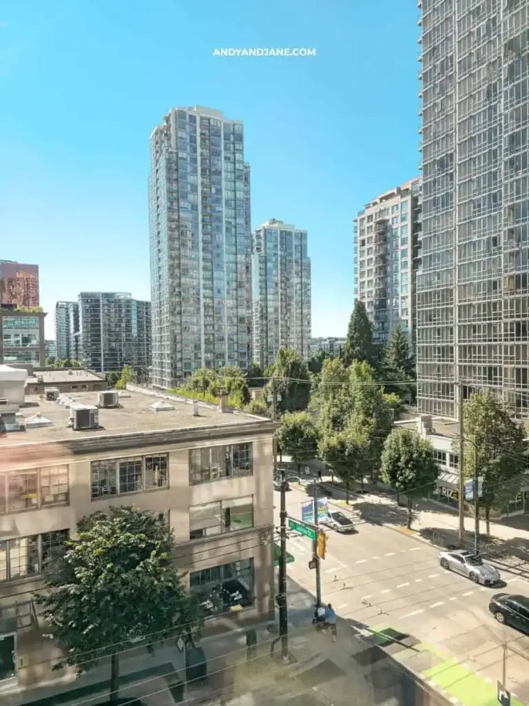 Cityscape with tall glass buildings under a clear blue sky. A tree-lined street runs below, cars are parked, and shadows cast a sunny, vibrant feel.